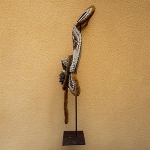 May include: A wooden mask with a long, flowing tail made of shells and fur. The mask is painted in black, white, and red, and has a human-like face. The mask is mounted on a black metal stand.