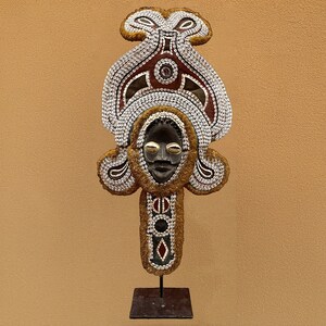 May include: A wooden mask with a human face, decorated with shells and beads. The mask is mounted on a black metal stand.