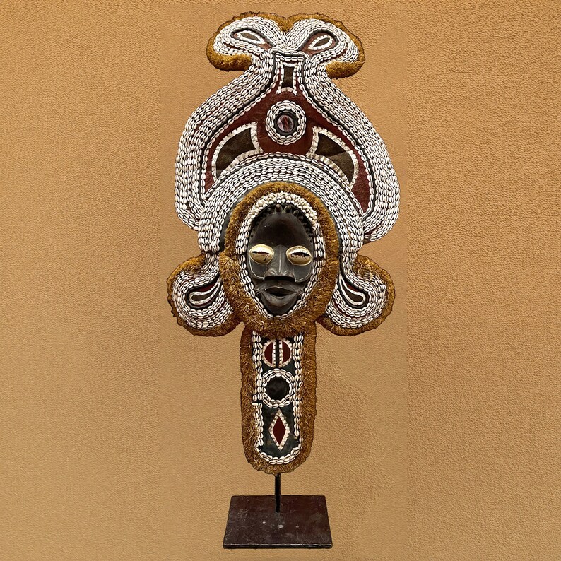May include: A wooden mask with a human face, decorated with shells and beads. The mask is mounted on a black metal stand.