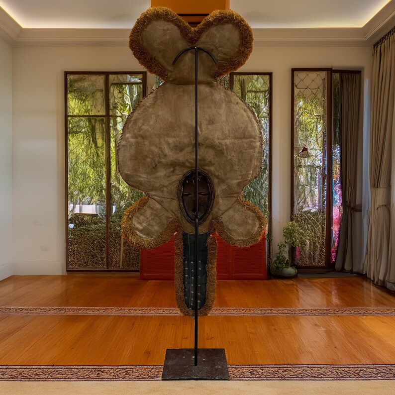 May include: A large, brown, fringed, fabric sculpture with a wooden mask in the center, mounted on a black metal stand. The sculpture is in a room with large windows and wooden floors.