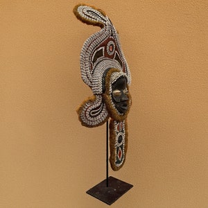 May include: A wooden mask with a human face, decorated with white and brown shells, red and white beads, and brown fur. The mask is mounted on a black metal stand.