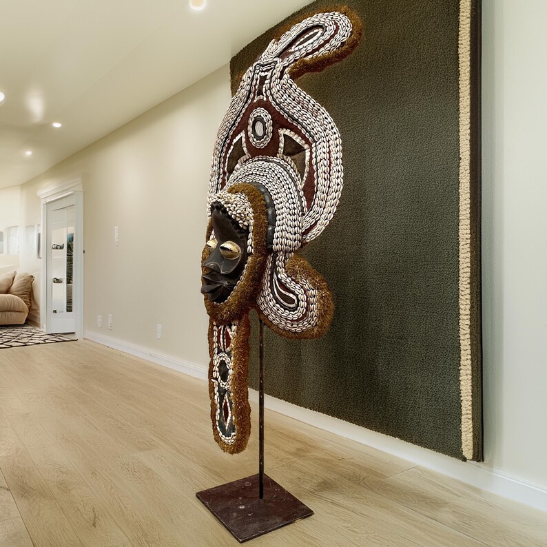 May include: A large wooden mask with intricate details and a brown, white, and gold color scheme. The mask is mounted on a metal stand and has a decorative fringe.