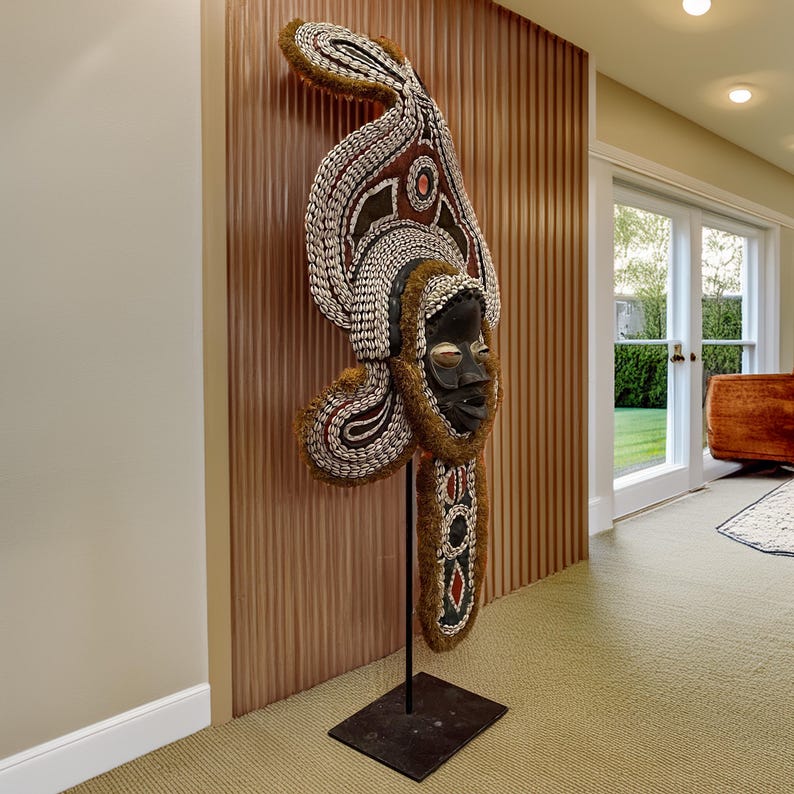 May include: A large, intricately carved wooden mask with a human face, adorned with shells and beads. The mask is mounted on a black stand and is displayed against a wood-paneled wall.