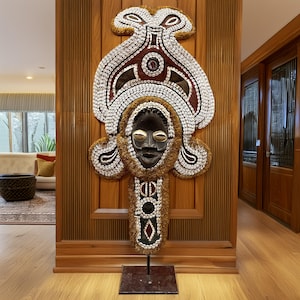 May include: A large, intricately decorated wooden mask with a human face, adorned with shells, beads, and feathers. The mask is mounted on a black stand and is displayed against a wooden wall.