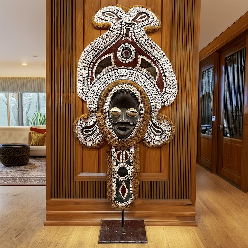 May include: A large, intricately decorated wooden mask with a human face, adorned with shells, beads, and feathers. The mask is mounted on a black stand and is displayed against a wooden wall.