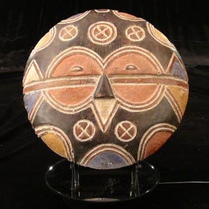 May include: A circular wooden mask featuring a carved face with two large circular eyes, a triangular nose, and a horizontal line across the center. The surface is decorated with geometric patterns in shades of brown, orange, blue, and white.