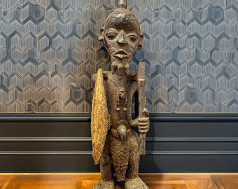 May include: A dark wood sculpture of a standing figure with a spear and shield. The figure has a carved face with a pointed hat and a detailed design on the chest and legs. The sculpture is standing on a wooden floor in front of a patterned wall.