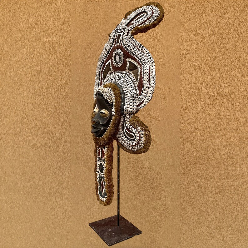 May include: A wooden mask with a decorative headdress made of shells and beads. The mask is brown and black with white and red accents. The headdress is white with brown and red accents. The mask is mounted on a black stand.