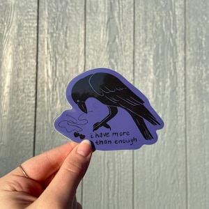 Puede incluir: Una pegatina morada con una ilustración de un cuervo negro mirando hacia abajo a una cadena de dos cerezas en forma de corazón. El texto dice "i have more than enough."