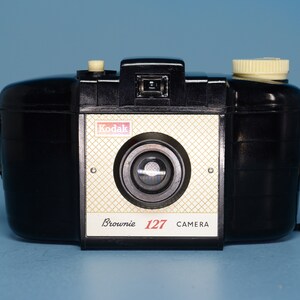 May include: A black Kodak Brownie 127 camera with a white label that reads "Kodak Brownie 127 CAMERA".