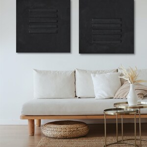 Puede incluir: Dos lienzos cuadrados negros con l&iacute;neas horizontales en relieve, colgados en una pared blanca sobre un sof&aacute; blanco con almohadas y un puf tejido. Dos mesas auxiliares de metal dorado con tapas de vidrio est&aacute;n frente al sof&aacute;.