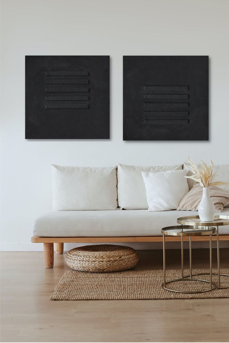 Puede incluir: Dos lienzos cuadrados negros con l&iacute;neas horizontales en relieve, colgados en una pared blanca sobre un sof&aacute; blanco con almohadas y un puf tejido. Dos mesas auxiliares de metal dorado con tapas de vidrio est&aacute;n frente al sof&aacute;.