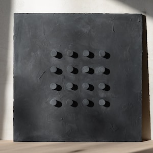 May include: A square, dark gray textured wall art piece with a grid of sixteen evenly spaced, cylindrical, raised elements. The artwork is set against a white wall with natural light casting shadows.