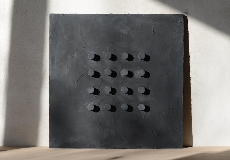 May include: A square, dark gray textured wall art piece with a grid of sixteen evenly spaced, cylindrical, raised elements. The artwork is set against a white wall with natural light casting shadows.