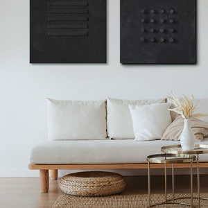 May include: Two black abstract wall art pieces with textured surfaces. One features horizontal lines, the other has a grid of circles. The art hangs above a light-colored sofa with white pillows, a woven footstool, and a jute rug.