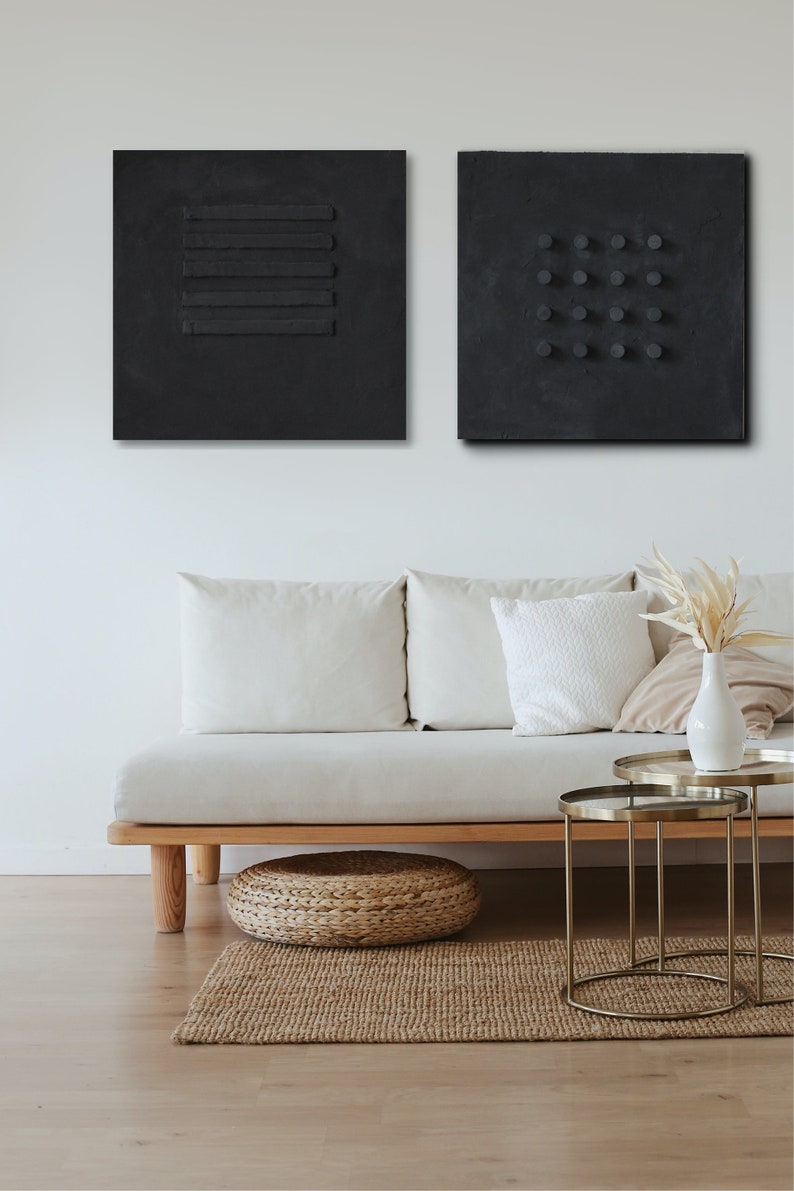 May include: Two black abstract wall art pieces with textured surfaces. One features horizontal lines, the other has a grid of circles. The art hangs above a light-colored sofa with white pillows, a woven footstool, and a jute rug.