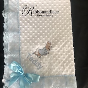 May include: White baby blanket with blue trim and lace edging. The blanket features an embroidered Peter Rabbit design and the name "Teddy". A blue satin bow is tied on the corner.