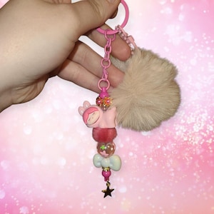 May include: A pink and white keychain with a sleeping cartoon character, a fluffy pom-pom, and a star charm.