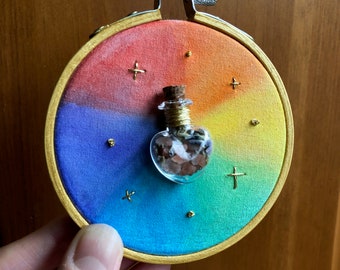 May include: A small, heart-shaped glass bottle filled with brown and white stones, with a gold wire around the neck, is displayed on a rainbow-coloured fabric stretched over a wooden embroidery hoop. The fabric is a gradient of red, orange, yellow, green, blue, and purple, with gold stars and crosses embroidered on it.