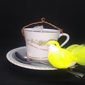 May include: A white teacup with a floral design and a copper wire handle sits on a light blue saucer with a spoon. A yellow bird decoration with green feathers is next to the cup. The background is black.