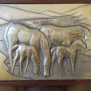 May include: A silver metal wall plaque depicting a group of four horses in a landscape setting. The horses are in a relief style and the plaque is framed in a dark wood frame. The plaque is signed "Mida's".