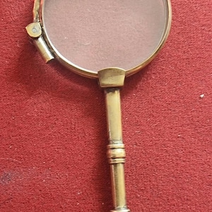 Puede incluir: Una lupa dorada con un mango plegable y un anillo en el extremo. La lupa está sobre una superficie roja.