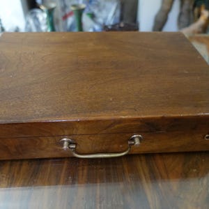 Peut inclure: Une boîte de rangement rectangulaire en bois avec une finition brun foncé et un grain de bois visible. La boîte comporte une poignée centrale en métal et deux petits loquets à crochet en métal sur le bord avant pour fermer le couvercle.