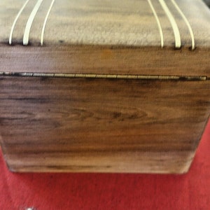 Op de afbeelding: Een houten doos met een scharnierend deksel. De doos is gemaakt van donker hout met een inleg van licht hout. Het deksel heeft een decoratief ontwerp met drie dunne lijnen van licht hout.