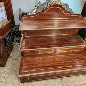 Op de afbeelding: Een houten displayplank met drie niveaus, voorzien van een donkere, gepolijste houtnerf en gouden metalen inlegaccenten. De unit bevat twee kleine lades op het middelste niveau en één grote lade aan de basis, bekroond met een decoratieve gouden metalen kuif.