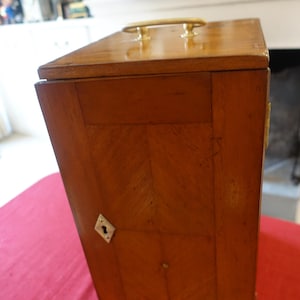 Puede incluir: Una caja de madera con una manija de latón y una cerradura. La caja tiene un patrón de veta de madera y está cerrada.