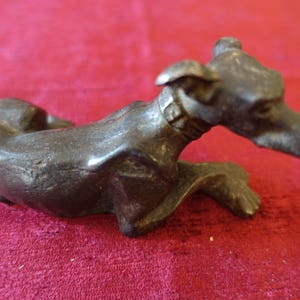 Op de afbeelding: Een bronzen sculptuur van een hond in rustpositie. De hond draagt een halsband en heeft een lange snuit en puntige oren. De sculptuur staat op een rode ondergrond.