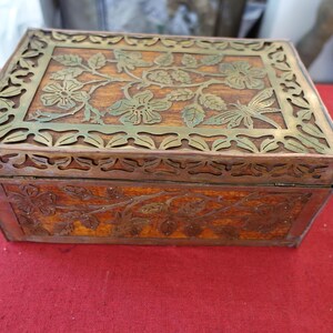 May include: A rectangular wooden box with a hinged lid. The lid and sides are adorned with floral and butterfly carvings. The top features a decorative brass overlay with cut-out floral patterns. The box is set on a red surface.