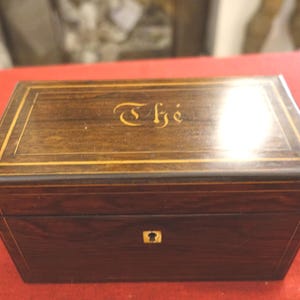 Peut inclure: Un coffret à thé rectangulaire en bois brun foncé, orné d'incrustations dorées et du mot "Thé" sur le couvercle. Le coffret présente une serrure sur le devant. Objet de collection.