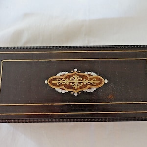 May include: A dark brown wooden box with a decorative inlay of gold and white. The inlay is in the shape of a stylized flower.