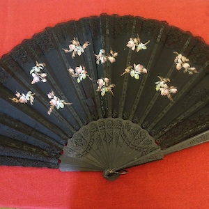 Può includere: Ventaglio nero con un design floreale. Il ventaglio presenta un bordo in pizzo nero ed è decorato con fiori rosa e bianchi dipinti. Il ventaglio è in legno e ha un manico ornato.