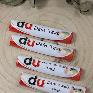 Könnte beinhalten: Vier Schokoriegel mit weißen Verpackungen und roten Akzenten. Die Verpackungen zeigen das Wort "du" in Schwarz und den Text "Dein Text" oder "Dein zweizeiliger Text". Die Riegel liegen auf einer Holzoberfläche.