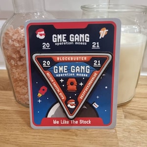 Könnte beinhalten: Ein blauer und roter Aufkleber mit dem Text "GME GANG" und "Operation moass" in Weiß. Der Aufkleber ist dreieckig mit einer Rakete, einer Person mit einem Weihnachtsmann-Hut und einem Pluszeichen auf jeder Seite. Der Text "BLOCKBUSTER" befindet sich über dem Dreieck und der Text "We Like The Stock" unter dem Dreieck.