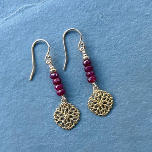 May include: A pair of gold-colored dangle earrings. Each earring features a strand of small, dark red beads and a gold filigree charm. The earrings hang from gold-colored ear wires.
