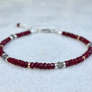 May include: A delicate bracelet featuring small red gemstone beads, silver beads, and a larger grey gemstone bead. The bracelet has a silver clasp.