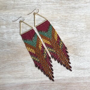 Puede incluir: Un par de pendientes largos con cuentas y un diseño geométrico. Los pendientes presentan una parte superior triangular de color dorado y una franja de cuentas en tonos burdeos, verde azulado, dorado y marrón. Los pendientes están diseñados para colgar y balancearse.