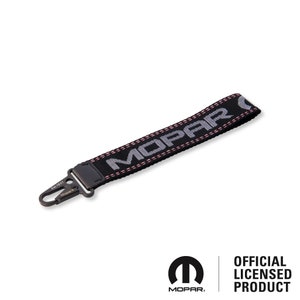 Könnte beinhalten: Schwarzer Schlüsselanhänger mit silbernem Mopar-Logo und rosa Nähten. Der Schlüsselanhänger hat einen Metallclip.