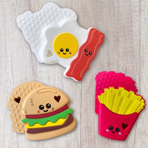 May include: A collection of colorful food-shaped silicone teethers. The set includes a fried egg, bacon, a hamburger, and a container of french fries, all with smiling faces and heart-shaped cutouts. The teethers are designed for babies.