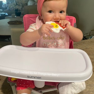 May include: A baby in a pink high chair with a white tray, wearing a pink headband and bib that says "babes who BRUNCH." The baby is chewing on a white, yellow, and red toy shaped like bacon and eggs.