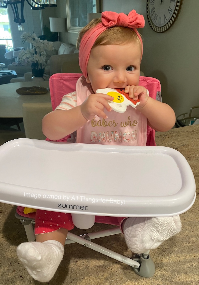 May include: A baby in a pink high chair with a white tray, wearing a pink headband and bib that says "babes who BRUNCH." The baby is chewing on a white, yellow, and red toy shaped like bacon and eggs.