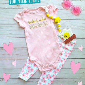 May include: A baby outfit set featuring a pink onesie with "babes who brunch" text, floral leggings, a pacifier clip with a star, and pink heart-shaped sunglasses. The set is displayed on a light wood background with pink heart cutouts.