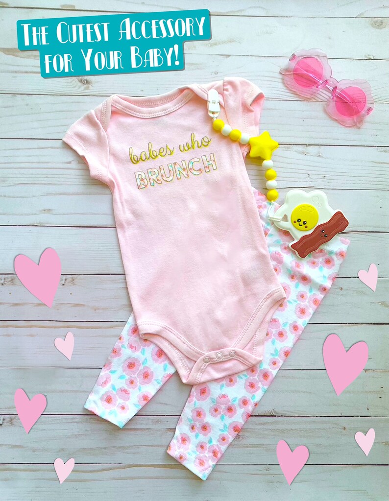 May include: A baby outfit set featuring a pink onesie with "babes who brunch" text, floral leggings, a pacifier clip with a star, and pink heart-shaped sunglasses. The set is displayed on a light wood background with pink heart cutouts.