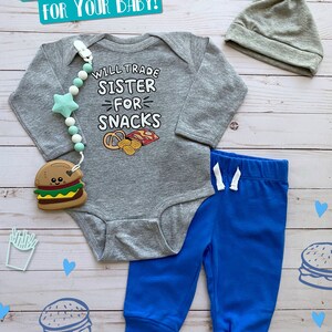 May include: A gray baby bodysuit with the text "WILL TRADE SISTER FOR SNACKS" and a pretzel and chip graphic. The set includes blue sweatpants, a gray knotted hat, and a burger teether. A pacifier clip with a star-shaped bead is also included.