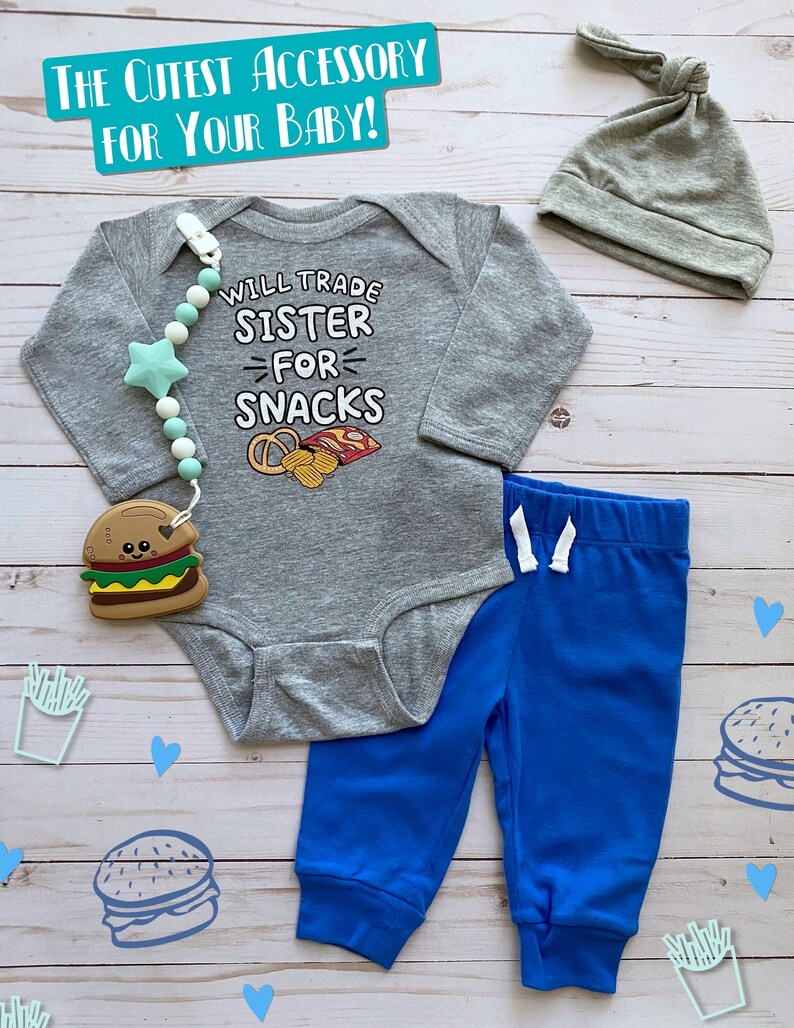 May include: A gray baby bodysuit with the text "WILL TRADE SISTER FOR SNACKS" and a pretzel and chip graphic. The set includes blue sweatpants, a gray knotted hat, and a burger teether. A pacifier clip with a star-shaped bead is also included.