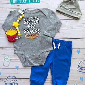 May include: A gray baby bodysuit with the text "WILL TRADE SISTER FOR SNACKS" and a matching blue pants set. The set includes a gray knotted hat and a pacifier clip with a star and french fry design.