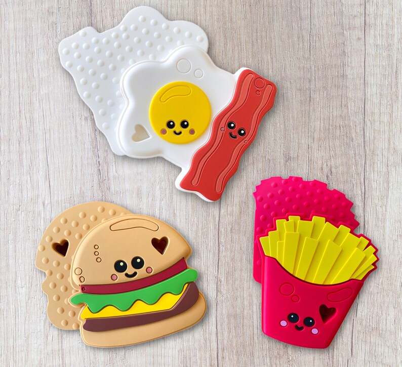 May include: A collection of colorful food-shaped silicone teethers. The set includes a fried egg, bacon, a hamburger, and a container of french fries, all with smiling faces and heart-shaped cutouts. The teethers are designed for babies.